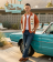 A man wearing a rust-colored and cream vintage bowling shirt featuring atomic-age starburst and oval embroidery, leaning against a classic turquoise car in front of a retro desert motel.