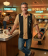 Gen X man wearing a gold and black vertical-paneled bowling shirt with embroidered bowling pins and starbursts, standing in a retro bowling alley lounge."