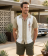 A man wearing a tan and cream vintage Hawaiian Guayabera shirt shirt with a geometric square and line embroidery on the chest, standing outdoors in front of a mid-century house and classic car.